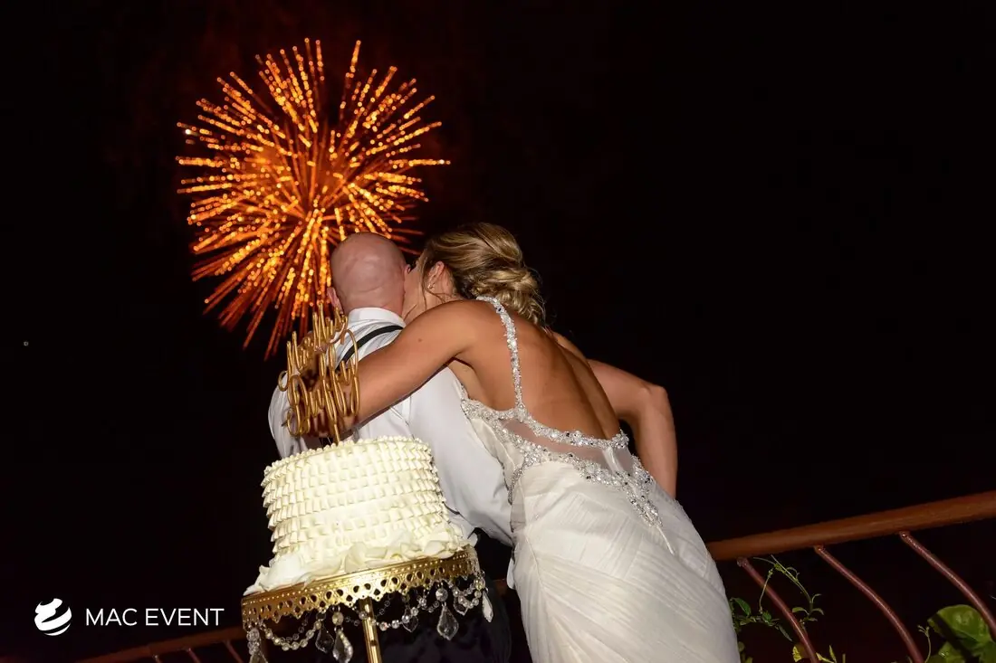 A bride and groom embrace as a golden firework display lights up the night sky behind them, with a wedding cake visible in the foreground.