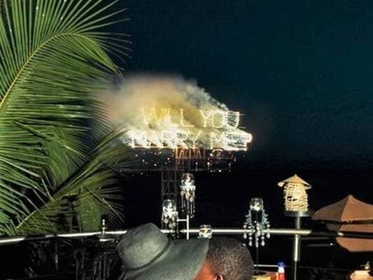 A marriage proposal display illuminated with fireworks spelling out "Will You Marry Me?" in a glowing, fiery sign against the night sky.